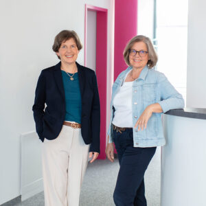 Two professional women in business casual attire stand in a bright office hallway, smiling at the camera.
