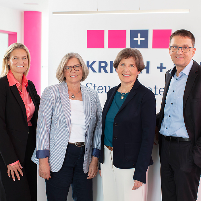Four professionals pose for a photo in a modern office, with a company sign featuring pink squares and a blue plus symbol in the background.