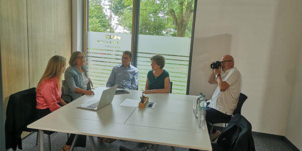 Four colleagues sit around a white conference table while a photographer on the right snaps a photo in a modern meeting room with large windows and greenery outside.