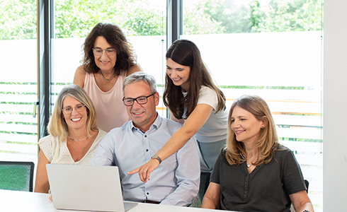 Five colleagues gather around a laptop in a bright office, with one person pointing at the screen as they discuss.