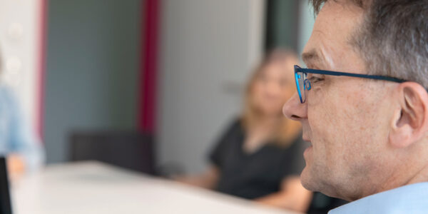 Close-up profile of a man wearing blue-framed glasses in a blurred office or meeting setting.
